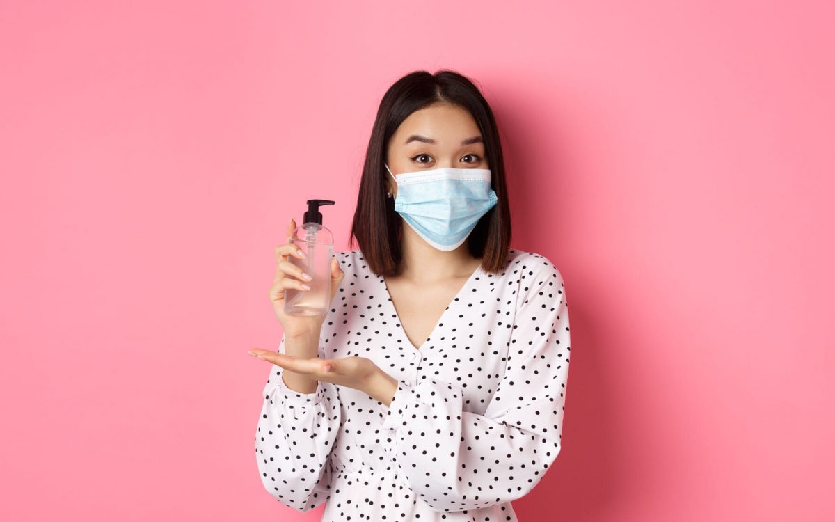 Covid-19, pandemic and lifestyle concept. Beautiful korean woman in dress and medical mask showing hand sanitizer, recommending antiseptic, standing over pink background.